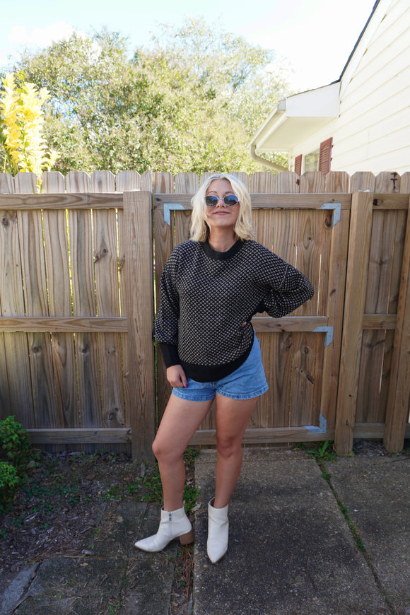 Person wearing a black sweater, denim shorts, and white boots standing in front of a wooden fence.