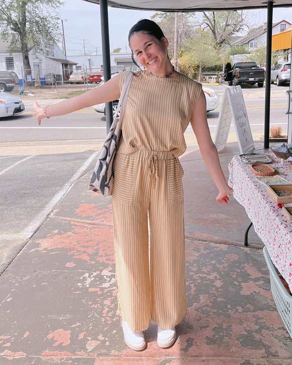 Woman in a beige jumpsuit standing on a sidewalk with a street and storefronts in the background.