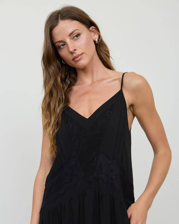 A woman modeling a black spaghetti-strap slip dress with lace detailing on the bodice, photographed against a plain light gray studio background.