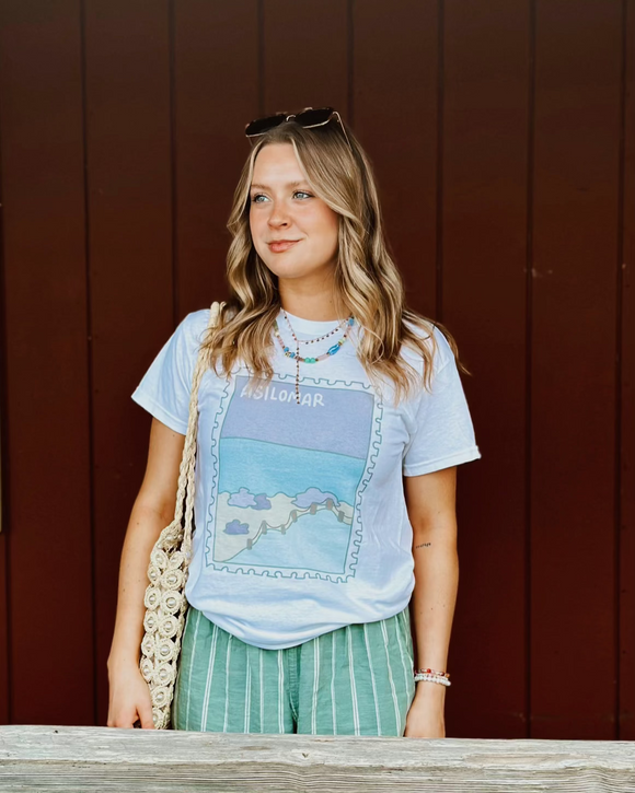 Woman wearing a graphic t-shirt and green pants standing against a red-brown wall.