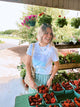 Woman shopping for strawberries at an outdoor market with flowers and plants in the background.
