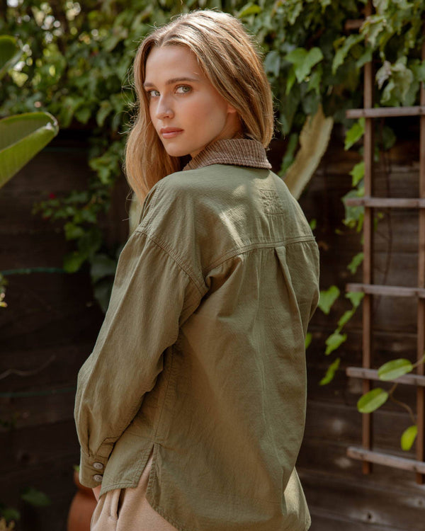 Woman wearing a green jacket standing outdoors with greenery in the background