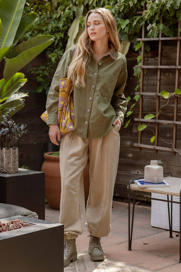 Woman standing outdoors in a garden setting wearing an olive green jacket and beige pants.