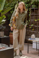 Woman standing outdoors in a garden setting wearing an olive green jacket and beige pants.