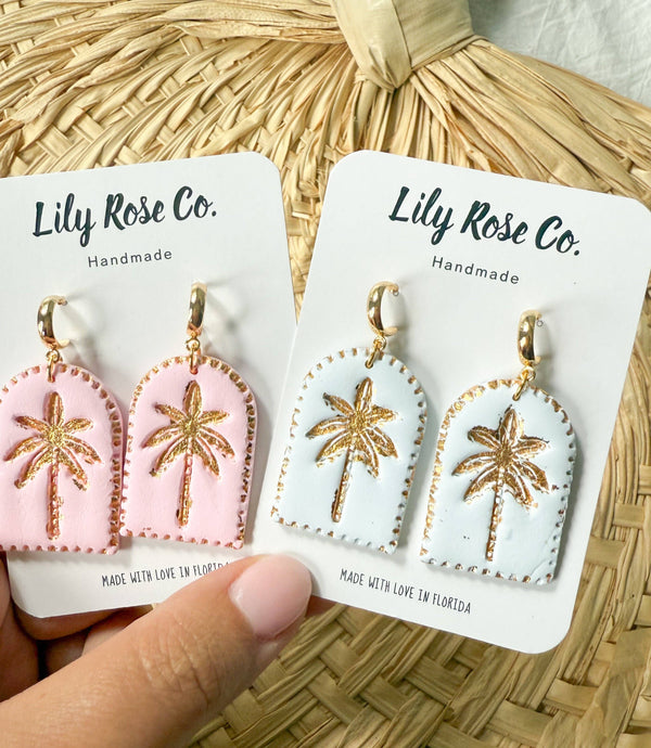 Pair of earrings with palm tree design on Lily Rose Co. cards, held against a woven background.