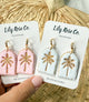 Pair of earrings with palm tree design on Lily Rose Co. cards, held against a woven background.