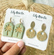 Two pairs of earrings on Lily Rose Co. cards held by a hand against a woven background.