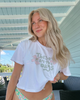Woman wearing a white t-shirt with a floral design and text, standing on a deck.