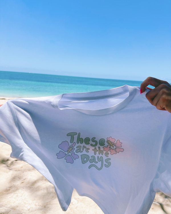 White t-shirt with 'These are the days' text and floral designs held by a person on a beach.