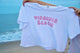 White t-shirt with 'Virginia Beach' printed in pink held by a person on a beach.