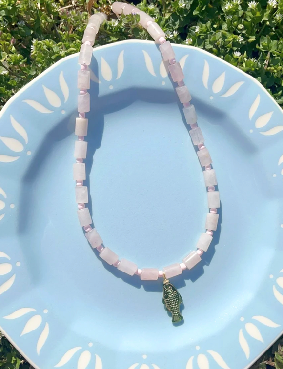 Necklace with pink beads and a green fish charm on a blue plate with white leaf patterns.