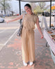 Woman in a beige jumpsuit standing on a sidewalk with a street and storefronts in the background.