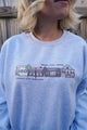 Light grey sweatshirt with a graphic design of buildings and text on a wooden background