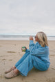 Person sitting on a beach holding a coffee cup, wearing a blue outfit.