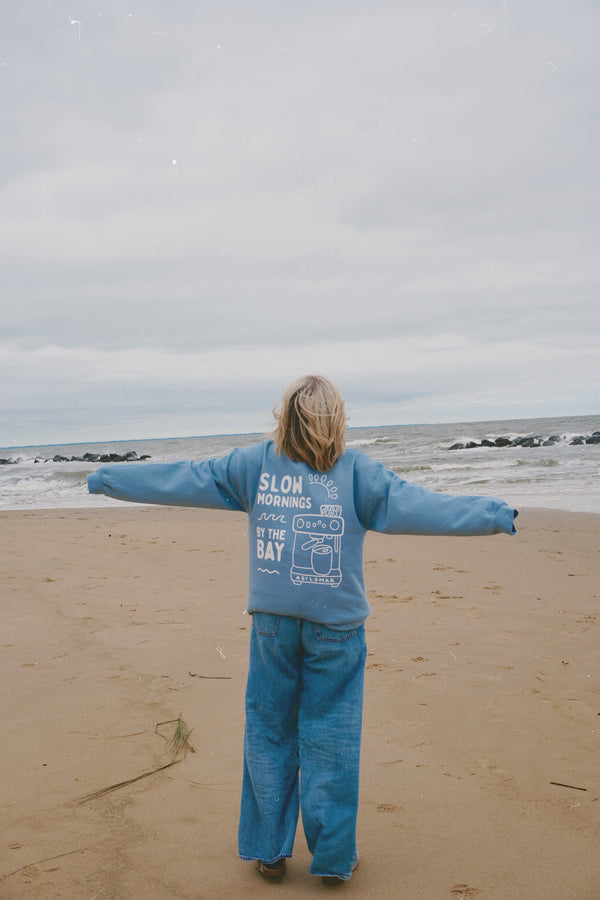 Person standing on a beach with arms outstretched, wearing a blue outfit with text.