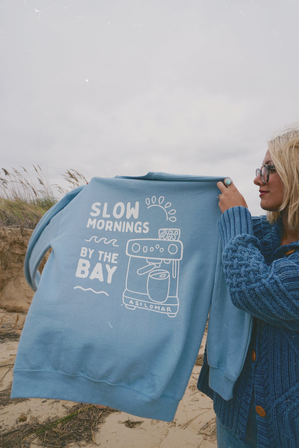 Person holding a blue sweatshirt with 'Slow mornings by the Bay' text on a beach.