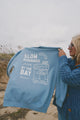 Person holding a blue sweatshirt with 'Slow mornings by the Bay' text on a beach.
