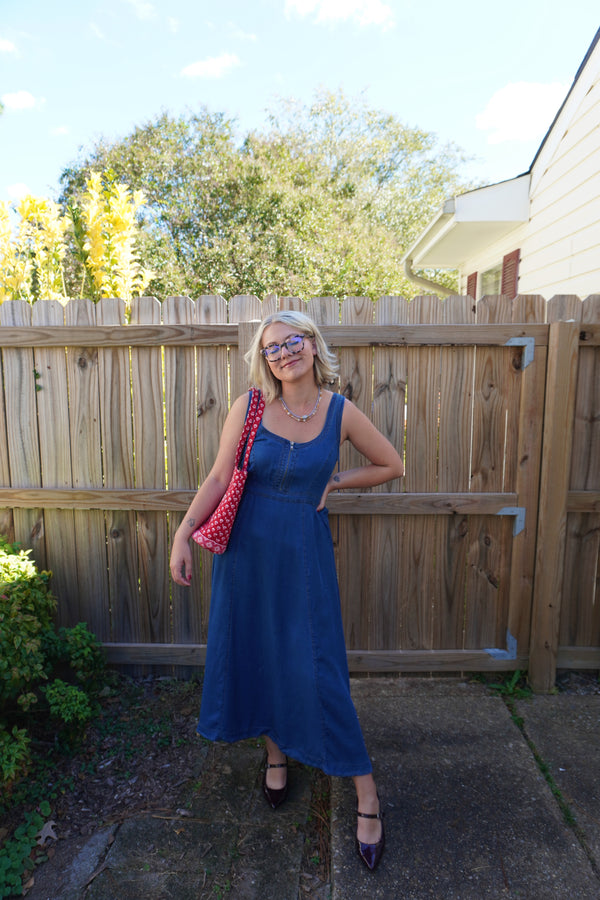 Woman in a blue dress standing outdoors with a wooden fence and trees in the background