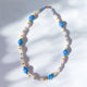 A beaded necklace featuring a mix of pastel and gold beads, with a prominent evil eye design, displayed against a shadowed background.