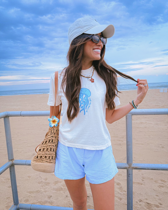 Woman at the beach wearing a white t-shirt with a jellyfish design, light blue shorts, and a white cap.