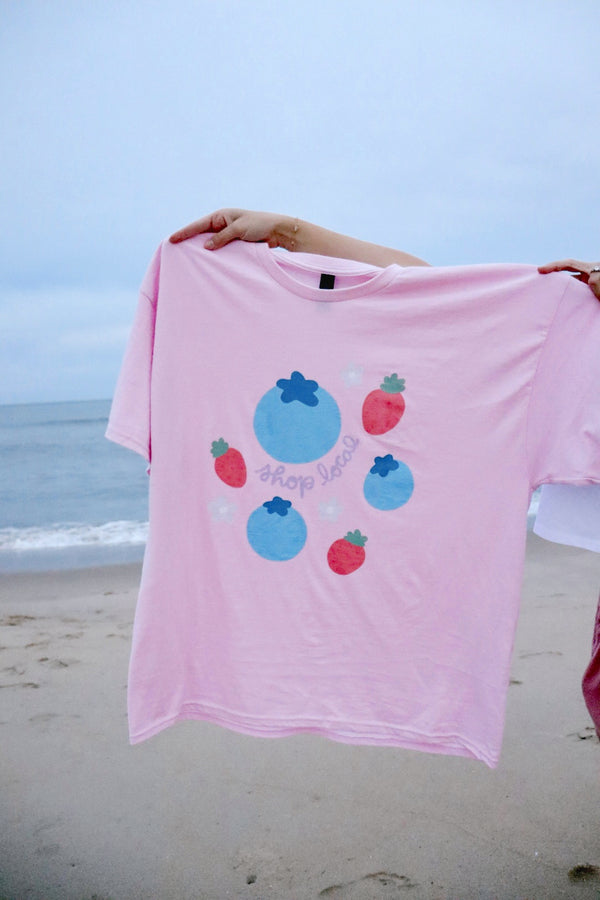 Pink t-shirt with colorful designs held by a person on a beach.