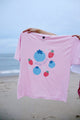 Pink t-shirt with colorful designs held by a person on a beach.