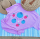 Purple shirt with colorful designs on a blue striped towel with hats in the background