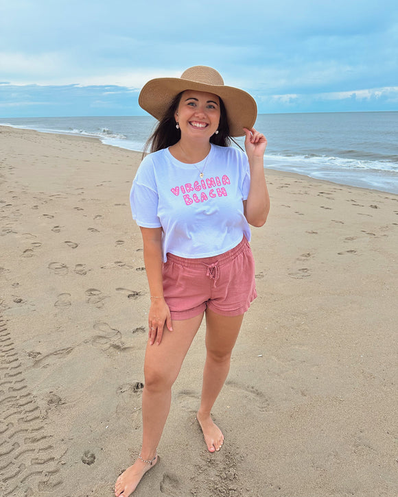 Woman on a beach wearing a white t-shirt with text and pink shorts, standing in front of the ocean.