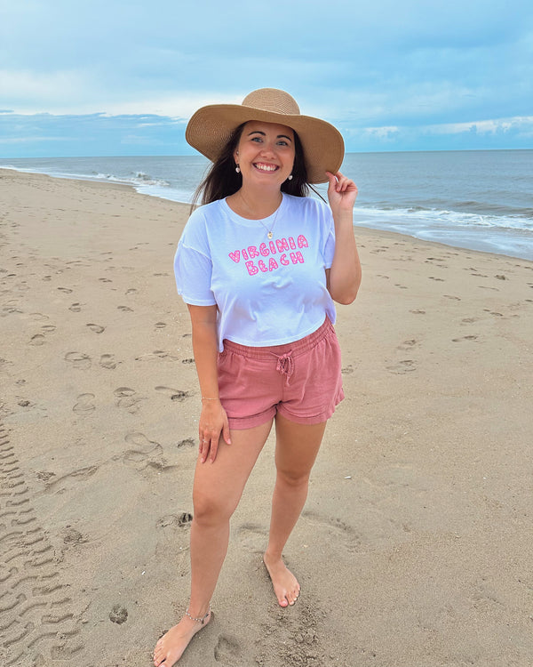 Woman on a beach wearing a white t-shirt with text and pink shorts, standing in front of the ocean.