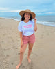 Woman on a beach wearing a white t-shirt with text and pink shorts, standing in front of the ocean.
